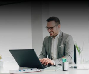 A man in a gray suit and glasses, resembling many successful CEOs, sits at a white desk, smiling while typing on a laptop. Office supplies, papers, and a plant are on the desk. The background is softly blurred.