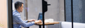 A man in a blue shirt works on a laptop at a small table in a modern glass office booth, possibly discussing hiring risks or building his sales team. Teapots and cups sit on the table beside him.