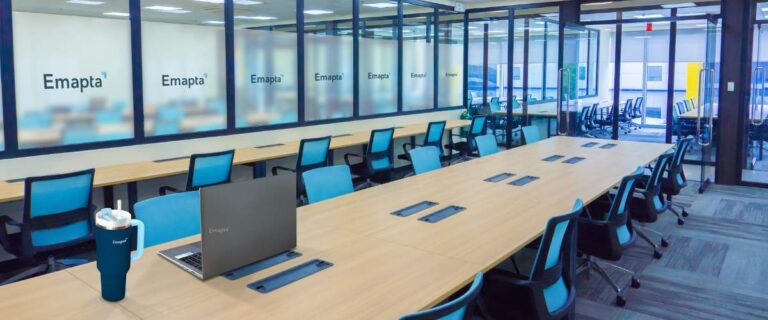 Modern conference room with long wooden tables, blue office chairs, and glass walls labeled Empata—perfect for teams deciding whether to hire freelancers or outsource to an agency. A laptop and tumbler rest on the table; another meeting room is visible through the glass.