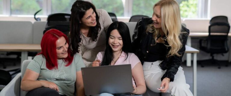 Four women sit and stand around a laptop in a bright office space, smiling and looking at the screen together, appearing engaged and happy as they discuss types of outsourcing for business.