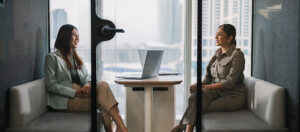 Two women sit across from each other in a modern office meeting room, each on a sofa bench, with a laptop open on the table between them. Large windows show a cityscape in the background.