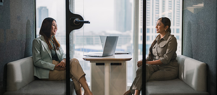 Two women sit across from each other in a modern office meeting room, each on a sofa bench, with a laptop open on the table between them. Large windows show a cityscape in the background.