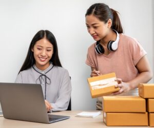 Two women working together at a desk; one is using a laptop and smiling, while the other, wearing headphones around her neck, is holding a small package—an eCommerce team managing orders with several cardboard boxes stacked nearby.