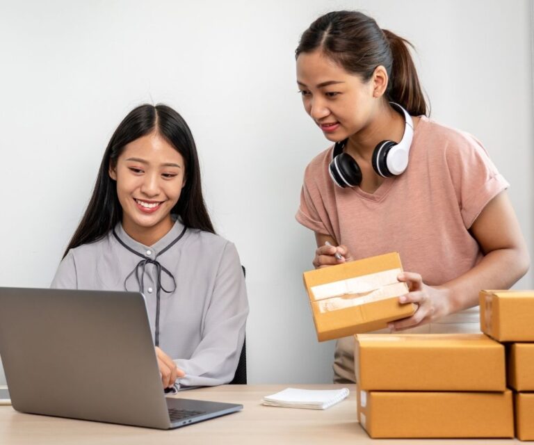 Two women working together at a desk; one is using a laptop and smiling, while the other, wearing headphones around her neck, is holding a small package—an eCommerce team managing orders with several cardboard boxes stacked nearby.