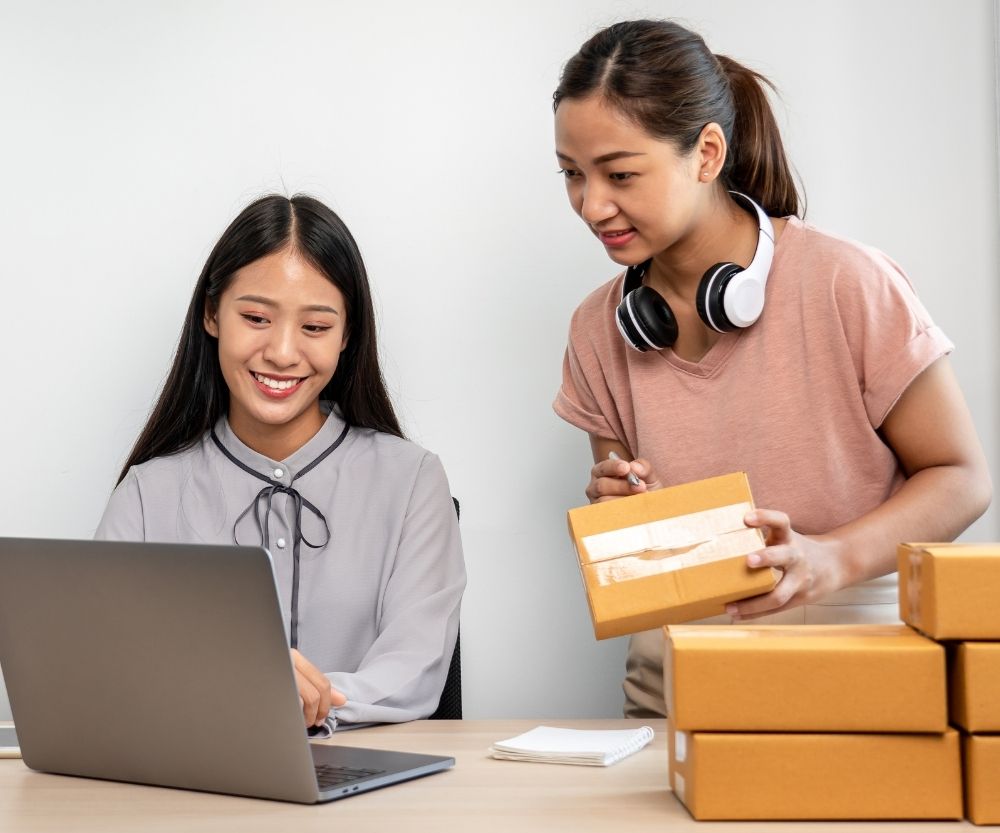 Two women working together at a desk; one is using a laptop and smiling, while the other, wearing headphones around her neck, is holding a small package—an eCommerce team managing orders with several cardboard boxes stacked nearby.