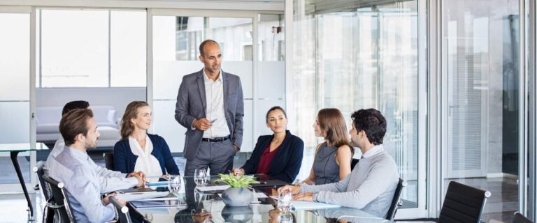Six professionals are seated around a glass conference table, attentively listening to a man discussing supply chain solutions in a modern office with large windows.