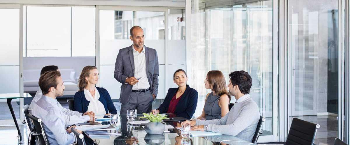 Six professionals are seated around a glass conference table, attentively listening to a man discussing supply chain solutions in a modern office with large windows.