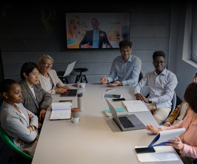 A diverse group of seven people sit around a meeting table with laptops, notebooks, and coffee cups, discussing supply chain nearshoring as a person joins the conversation via video call on a screen at the front of the room.