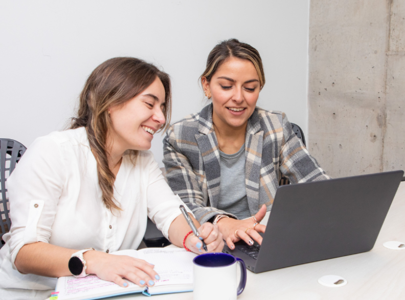 Two women sitting at a table, one writing in a notebook and the other using a laptop. Both are smiling and appear to be collaborating, showcasing how to build relationships in direct marketing. A coffee mug and a pen are also on the table.