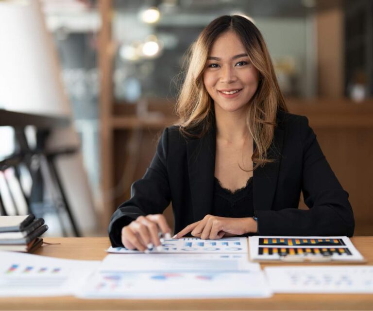 A Filipino accountant in a black blazer sits at a desk with charts and graphs, smiling at the camera in a modern office setting—ready to be your strategic partner.