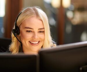 A smiling woman with blonde hair wearing a headset sits at a desk, looking at a computer monitor in an office setting—reflecting the modern nearshoring trends popular among European businesses by 2026.