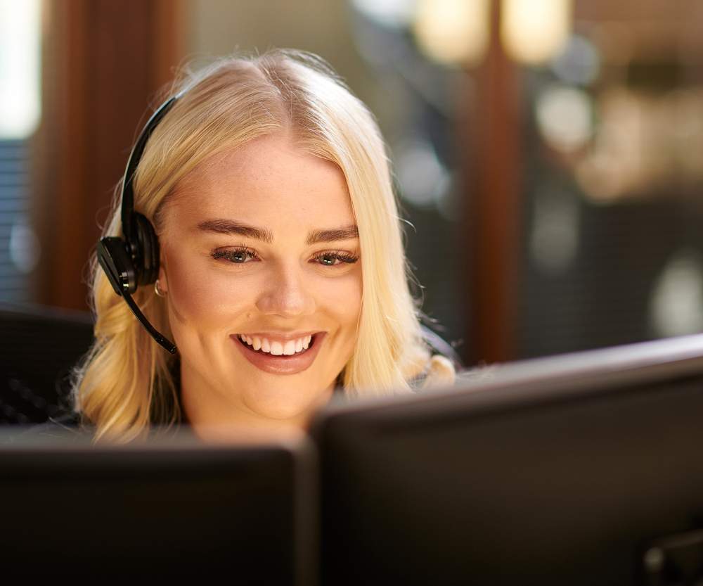 A smiling woman with blonde hair wearing a headset sits at a desk, looking at a computer monitor in an office setting—reflecting the modern nearshoring trends popular among European businesses by 2026.