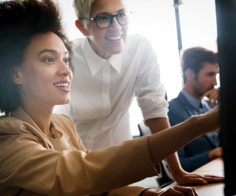 Two women in an office smile and look at a computer screen together, while a man works at his desk in the background. The atmosphere is collaborative and positive—ideal for North American businesses exploring nearshoring opportunities.