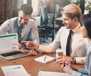 Three people in business attire sit around a table. One man points at charts and graphs on a laptop screen, highlighting outsourcing benefits, while the others watch and smile. Papers and a coffee cup are on the table in a bright office.