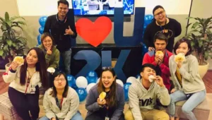 A group of nine smiling people pose indoors with snacks, sitting and standing in front of blue balloons and a large “I ♥ U 2K” sign. Capturing the warmth of life in the Philippines, green plants decorate both sides in the background.