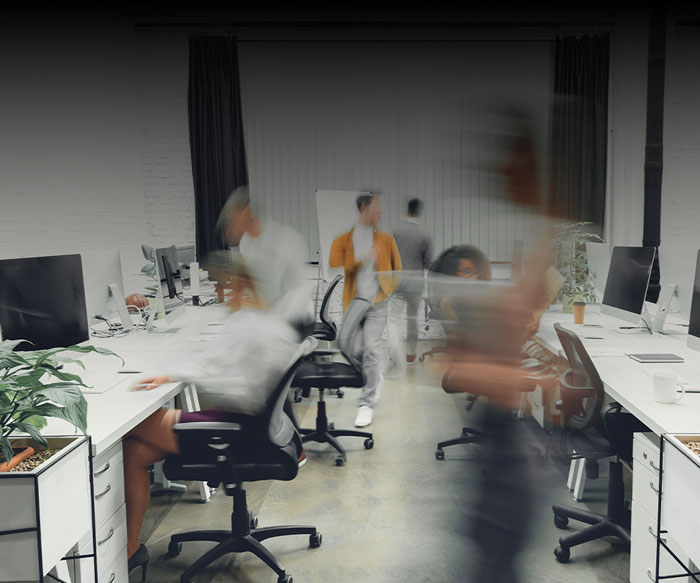 A modern office with several people moving around, creating a blurred effect. Desks with computers, chairs, and indoor plants set the scene as a hub of activity—challenging common outsourcing myths in a dynamic workspace.