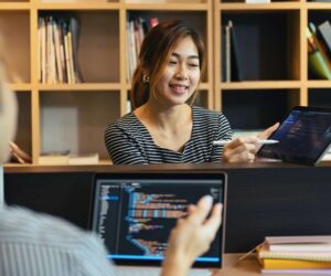 Two people sit at a desk with laptops displaying code. One person, smiling, gestures to her laptop screen while discussing ways to boost efficiency in IT operations. Bookshelves are in the background.