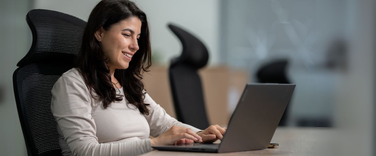 A woman with long dark hair sits at a desk in an office, smiling as she types on a laptop. Focused and content in her light-colored top, she appears confident while tackling common outsourcing problems with effective solutions.