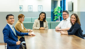 Five people sit around a wooden conference table in a modern office, smiling at the camera. The background features light walls, framed artwork, and green plants—ideal for discussing outsourcing tips or planning to outsource overseas.