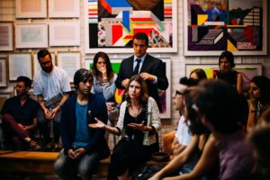 A woman gestures while speaking to a group of Colombian workers in an art gallery, with colorful abstract paintings on the wall behind them. The audience listens attentively during a culture training session, some sitting and some standing.