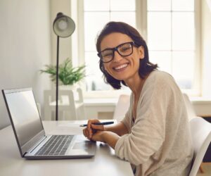 A woman with glasses smiles at the camera while working remotely from Colombia, sitting at a desk with a laptop and holding a pen. A plant and lamp are in the background by a sunny window, reflecting the rise of remote work in Colombia.