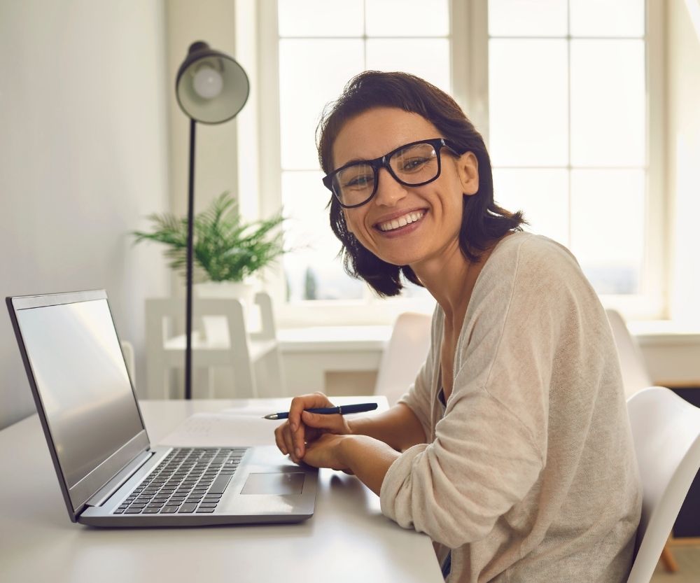 A woman with glasses smiles at the camera while working remotely from Colombia, sitting at a desk with a laptop and holding a pen. A plant and lamp are in the background by a sunny window, reflecting the rise of remote work in Colombia.