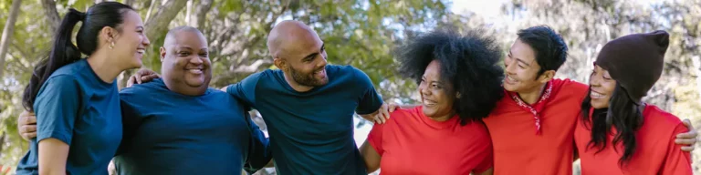 A diverse group of six adults stands outdoors, smiling and embracing each other in a line, wearing casual red and blue shirts—showcasing the power of camaraderie and connection vital for any successful employee engagement plan.
