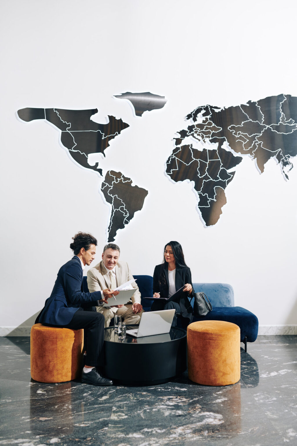 Three business professionals sit together in a modern office lounge, reviewing a 2025 Preparedness Guide. A large world map is displayed behind them, while two orange ottomans and a blue sofa surround a black coffee table.