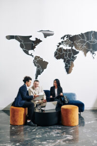 Three business professionals sit together in a modern office lounge, reviewing a 2025 Preparedness Guide. A large world map is displayed behind them, while two orange ottomans and a blue sofa surround a black coffee table.