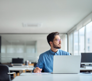 A man with glasses sits at a desk in a modern office in Latin America, looking thoughtfully to the side while holding a pen. A laptop is open before him, and the workspace is bright with large windows—reflecting an emerging IT hub.