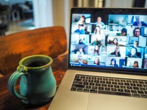 A green ceramic mug sits on a wooden table next to a laptop showing a video conference with many participants, as an outsourcing guide is discussed on the screen.