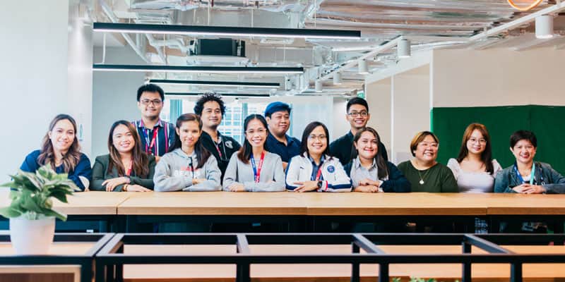 A group of thirteen people smiling and standing behind a long wooden table in a modern, well-lit office space highlights the collaborative spirit often seen in Philippines outsourcing. A potted plant is on the table in the foreground.