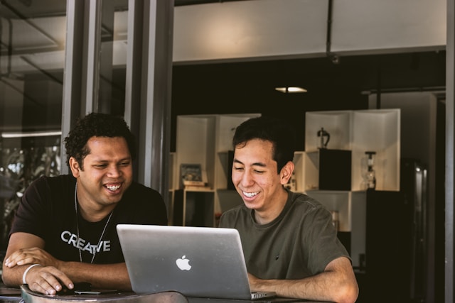 Two young men are sitting together at a table, smiling and looking at the screen of an open laptop in a modern indoor setting, showcasing the vibrant Colombia bilingual talent ideal for outsourcing.