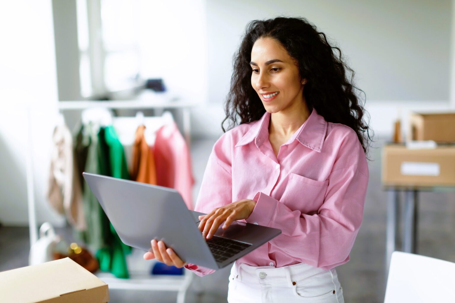 A woman with long curly hair, wearing a pink shirt and white pants, stands indoors using a laptop. Behind her are racks of colorful clothes and boxes, highlighting a busy workspace where e-Commerce outsourcing helps streamline operations.