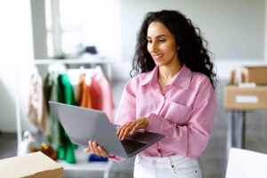 A woman with long curly hair, wearing a pink shirt and white pants, stands indoors using a laptop. Behind her are racks of colorful clothes and boxes, highlighting a busy workspace where e-Commerce outsourcing helps streamline operations.