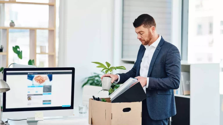 A man in a suit stands by a desk, packing office items such as a plant and folders into a cardboard box, capturing the impact of the Great Resignation. A computer monitor and office supplies are visible in the background.