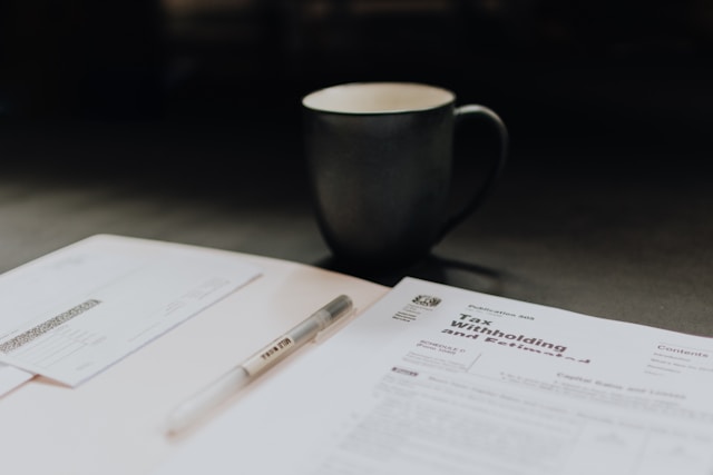 A ceramic mug sits on a dark surface behind a pen and paperwork, including a tax withholding form and an envelope, suggesting a workspace focused on managing finances or ensuring US remote work compliance.