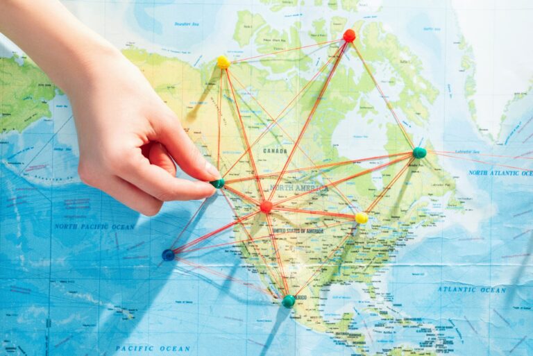 A hand places a pin on a map of North America, connecting colorful pins with string to illustrate a nearshoring network of business locations across the United States and Canada.