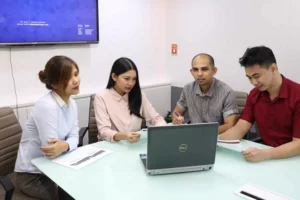 Four people sit around a table in an office, discussing documents. A laptop is open before them, and a large screen is mounted on the wall, as they explore the differences between outsourcing and offshoring during their meeting.