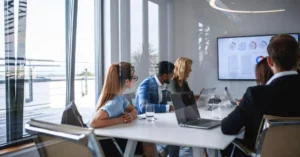 Four people sit around a conference table in a modern office, attentively watching a presentation on team migration strategies, with charts and graphs displayed on a large screen. Laptops and documents are spread across the table.