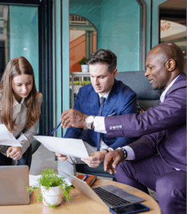 Three business professionals sit together at a table with laptops and documents, discussing paperwork in a modern office setting. They explore tech innovations to support colleagues with dyslexia as one man points at a sheet while the others listen intently.