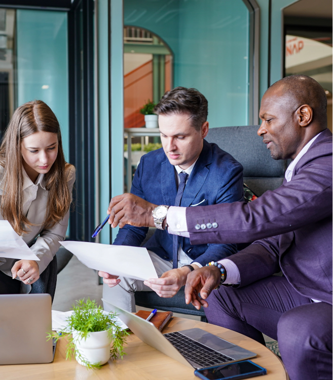 Three business professionals sit together at a table with laptops and documents, discussing paperwork in a modern office setting. They explore tech innovations to support colleagues with dyslexia as one man points at a sheet while the others listen intently.