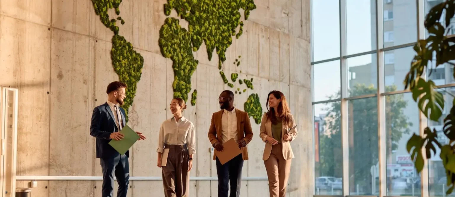 Four business professionals walk and talk in a modern office with large windows. Behind them, a world map made of green plants symbolizes sustainability, growth, and global connection inspired by their commitment to integrity.