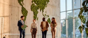 Four business professionals walk and talk in a modern office with large windows. Behind them, a world map made of green plants symbolizes sustainability, growth, and global connection inspired by their commitment to integrity.