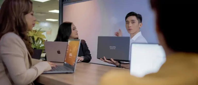 Four people sit around a table with laptops, engaged in a discussion about continuity strategy. One man holds up his laptop and gestures while speaking, as the others listen attentively in a modern office meeting room.
