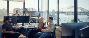 A group of people sit and talk around desks with computers in a modern office with large windows, overlooking a harbor and a bridge—an inspiring setting where Australian businesses discuss compliance and outsourcing on a sunny day.