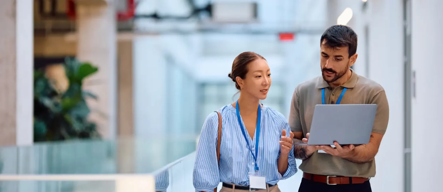 Two professionals wearing name badges and blue lanyards walk through a modern office hallway, discussing an outsourcing comparison on an open laptop. One person gestures while speaking, highlighting key points about Outsourcing in the Philippines and India.
