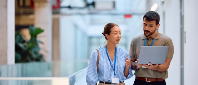 Two professionals wearing name badges and blue lanyards walk through a modern office hallway, discussing an outsourcing comparison on an open laptop. One person gestures while speaking, highlighting key points about Outsourcing in the Philippines and India.