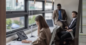 Three professionals are in a modern office by large windows. A woman works on a tablet, while two men dressed in business attire from Costa Rica and Colombia sit and talk in the background. Natural light fills the workspace.