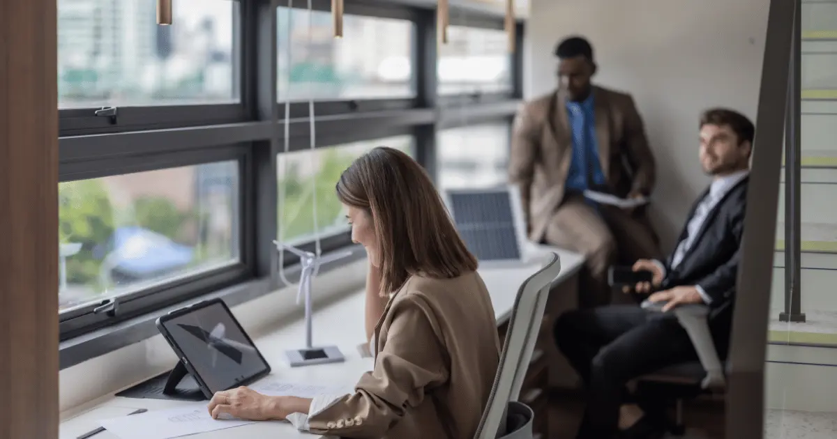 Three professionals are in a modern office by large windows. A woman works on a tablet, while two men dressed in business attire from Costa Rica and Colombia sit and talk in the background. Natural light fills the workspace.